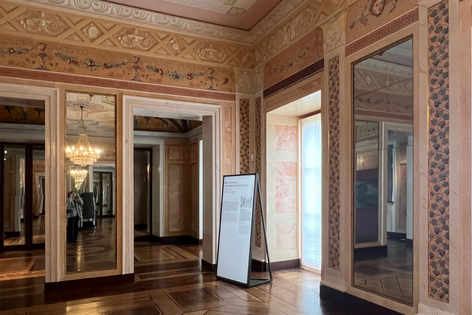 Blick in das Schloss Weimar. Ein Raum mit floralen Ornamenten und Holzboden. Zentral steht eine Informationstafel im Hochformat.